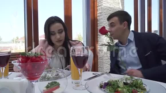 他在餐桌旁的餐馆里给了女孩一朵红玫瑰他们亲吻慢动作的股票录像视频的预览图