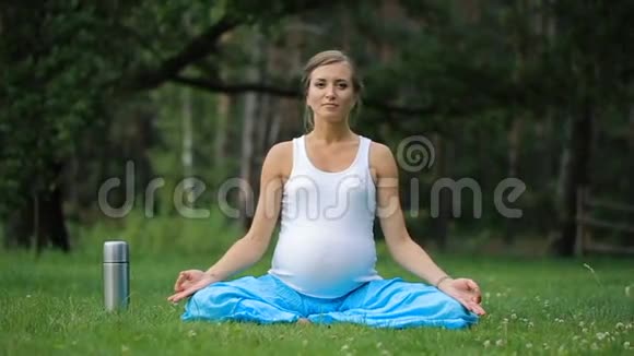 怀孕瑜伽在莲花的位置与热水瓶喝茶在公园的草地上户外健康的女人女人视频的预览图