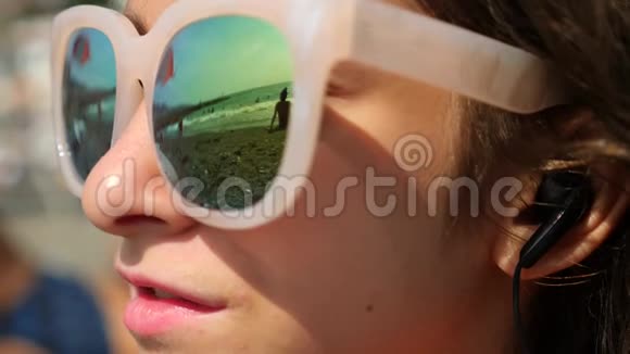 戴太阳镜的少女坐在海滩上用智能手机唱一首歌用耳机听她说话在里面视频的预览图