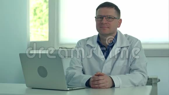 成熟的男医生坐在办公桌上手提电脑在他的诊所里看着相机视频的预览图