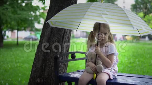 一个带着玩具兔子的小女孩坐在伞下的长凳上视频的预览图