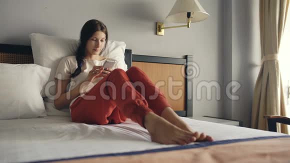 漂亮的金发女人躺在酒店房间的床上用智能手机微笑视频的预览图