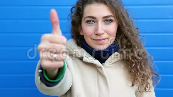 美丽的年轻女子伸出大拇指微笑倚在蓝色的墙上视频的预览图
