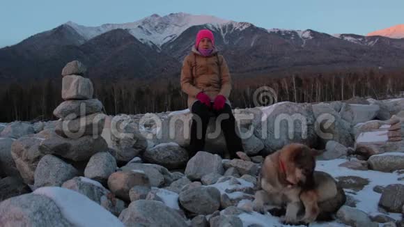 年轻女子和她的狗坐在山脚下的岩石上视频的预览图