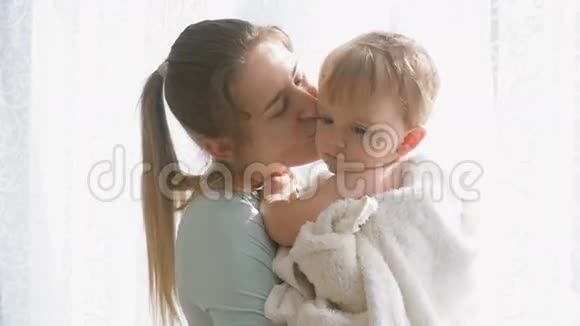 快乐的年轻母亲在大窗户上拥抱和亲吻她的小儿子的慢镜头视频的预览图