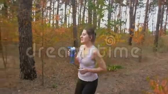 美丽苗条的女人在森林里奔跑从瓶子里喝水的慢镜头视频的预览图