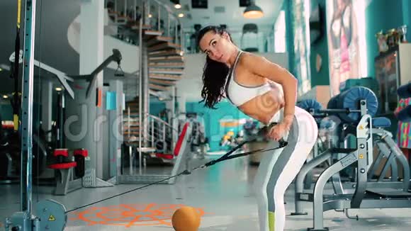女人用模拟器训练双手用弯曲的腿站在健身房里视频的预览图