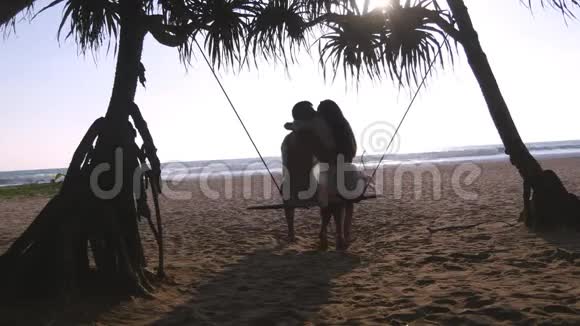年轻浪漫的情侣一起坐在热带海滩的秋千上拥抱恋爱中的伴侣放松和亲吻视频的预览图
