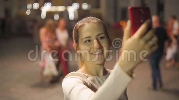 美丽的微笑女人在夜街的智能手机上自拍视频的预览图