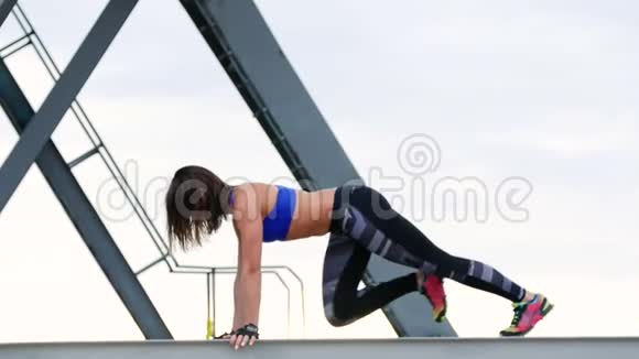 穿着蓝色上衣和条纹腿裤的运动女性进行力量练习她站在货运的金属横梁上视频的预览图