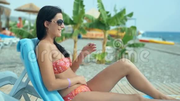 一位穿着泳衣在海滩上的躺椅上放松的年轻女子视频的预览图