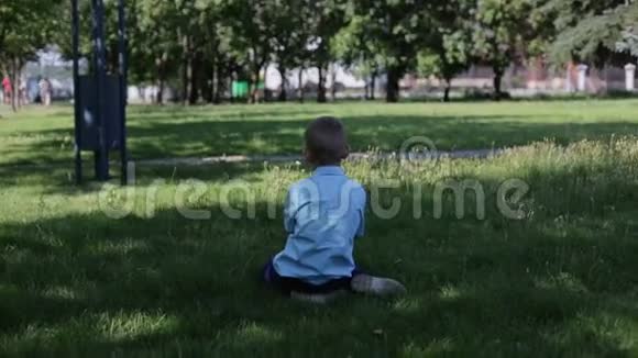 穿着蓝色衬衫的小悲伤男孩在公园里迷路了独自坐在草地上视频的预览图