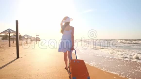 一个年轻的女人带着一个手提箱坐在海滩上一个年轻的女孩带着一个轮式手提箱沿着海滩散步一路走来视频的预览图