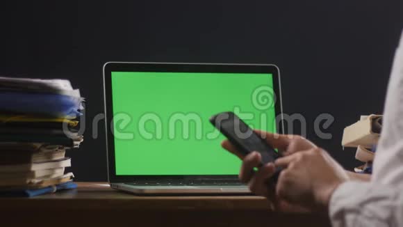 人类正在使用智能手机他的笔记本电脑上有一个绿色屏幕或一个色度在他的桌面上商人在这里工作视频的预览图