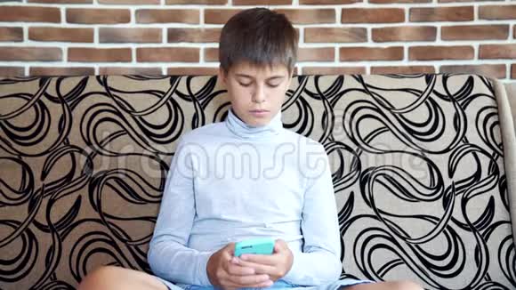 男孩坐在房间里的沙发上用智能手机现代技术在儿童生活中的概念视频的预览图