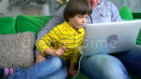 父子使用笔记本电脑英俊的父亲看着电脑他和非常可爱的儿子在家里的沙发上背景视频的预览图