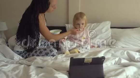 年轻漂亮的女人一边看平板电脑一边喂小女孩粥视频的预览图
