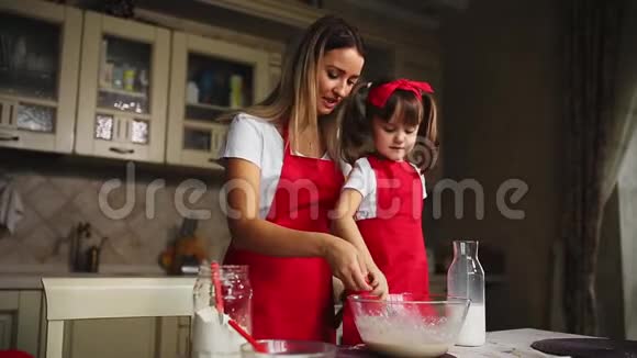 美丽的年轻母亲帮助她的小女儿一起用红色围裙做蛋糕把面粉倒入碗里打一打视频的预览图