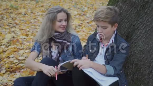 儿子和妈妈一起做作业坐在公园的秋叶旁视频的预览图