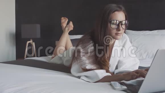 女商人戴着眼镜手提电脑躺在酒店的床上电脑上的智能手机在线商务视频的预览图