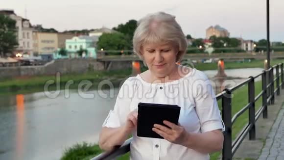 傍晚时分在海滨旧城河桥附近使用平板电脑的老年妇女视频的预览图