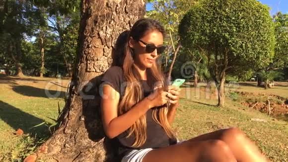 女孩坐在公园里的树附近用智能手机拍摄自拍照片慢速高清镜头视频的预览图
