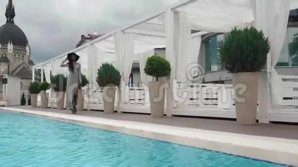 慢动作优雅的非裔美国女性模特穿着鸡尾酒条纹连衣裙和黑色帽子在豪华游泳池散步视频的预览图