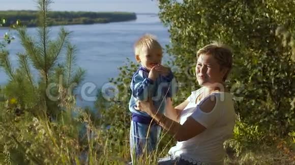 阳光明媚的夏日幸福的母亲和她的小儿子坐在河边的画像视频的预览图