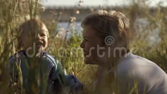 年轻漂亮的母亲和儿子在河边美丽的田野里玩视频的预览图