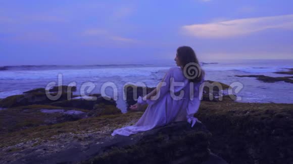 美丽的年轻女性穿着长裙坐在夕阳西下的巴厘岛海滩上的岩石上美丽的景色视频的预览图