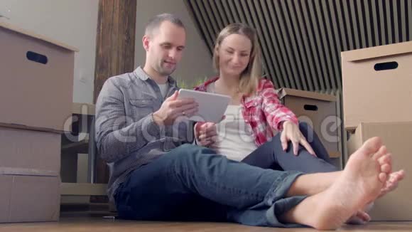 年轻夫妇坐在平板电脑的地板上为他们的新公寓选择家具视频的预览图