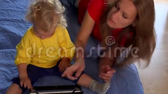 保姆教小蹒跚学步的女孩使用平板电脑坐在床上视频的预览图
