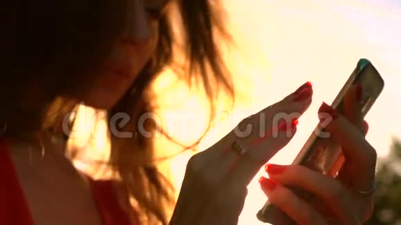 美丽的女孩用红色指甲油使用她的智能手机在公园温暖的日落颜色慢速视频视频的预览图