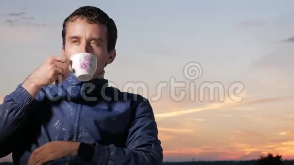 日落时一个年轻人从杯子里喝一杯热饮料后面是美丽的天空视频的预览图