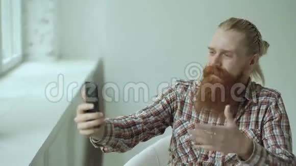 一位留胡子的年轻时髦商人坐在椅子上在智能手机上聊天视频视频的预览图