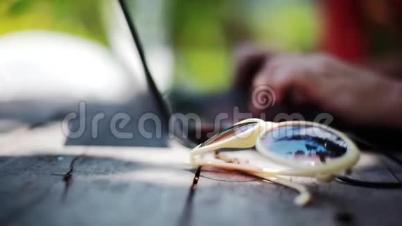 在热带小岛上一双手拿着笔记本电脑工作背景模糊太阳眼镜旁边视频的预览图