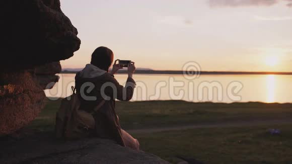 年轻的旅游女性背包客在日落时分在岩石上徒步旅行后在智能手机摄像头上拍摄风景视频的预览图