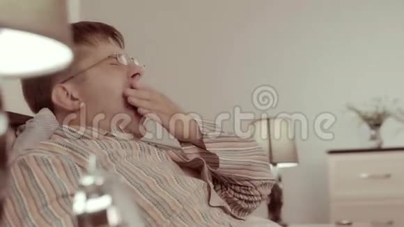 打哈欠的年轻人把眼镜放在床头柜上睡觉视频的预览图
