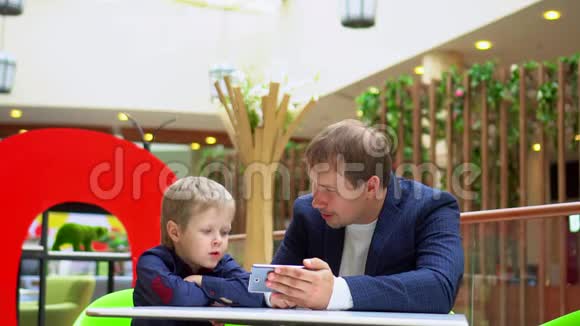 父亲和儿子坐在贸易商场的一张小桌子旁父亲带着儿子在电话里看视频母亲视频的预览图
