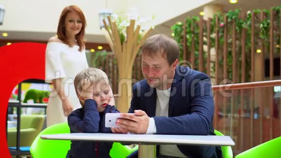 他父亲和儿子在电话里看视频母亲从后面走近一家人年轻的幸福家庭购物视频的预览图