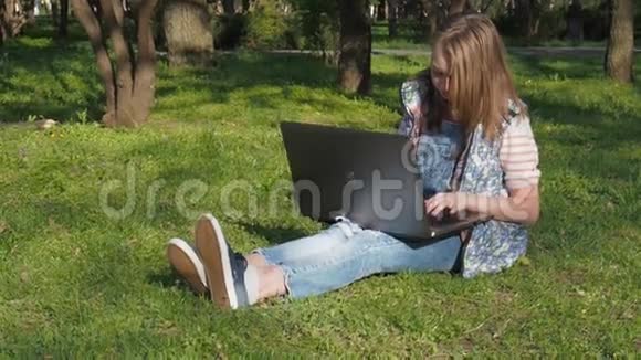 孩子在笔记本电脑上工作一个戴着太阳镜的十几岁的女孩坐在公园的草地上女孩阳光明媚的户外日洛杉矶视频的预览图