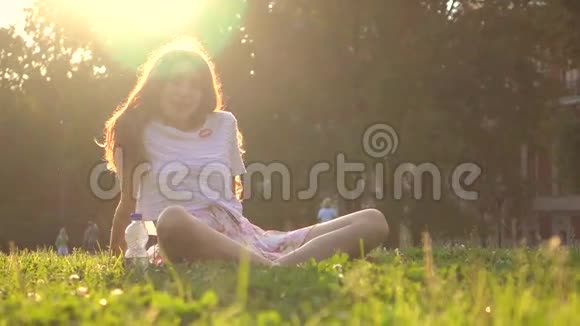 美丽的黑发女孩坐在公园的草地上在明亮的阳光下放松4K镜头视频的预览图