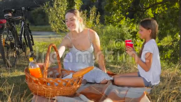 快乐的年轻妈妈和女儿坐在树下喝橙汁视频的预览图