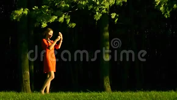 美丽的黑发女孩穿着红色连衣裙在公园里用手机拍照慢速视频视频的预览图