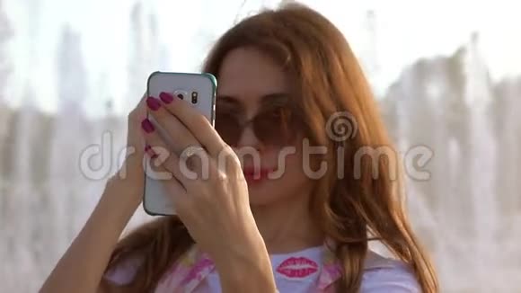 美丽的黑发女孩戴着太阳镜用手机在大公园喷泉附近自拍4K视频视频的预览图