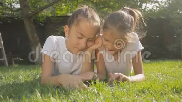 两个微笑的女孩在草地上放松玩数字平板电脑视频的预览图
