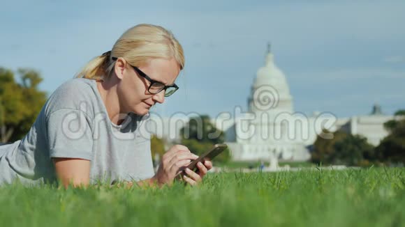 一位女士躺在华盛顿国会大厦背景下的草坪上使用智能手机旅游学习和视频的预览图