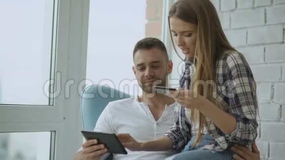 年轻的情侣们用数码平板电脑上网购物坐在现代阁楼公寓的阳台上视频的预览图