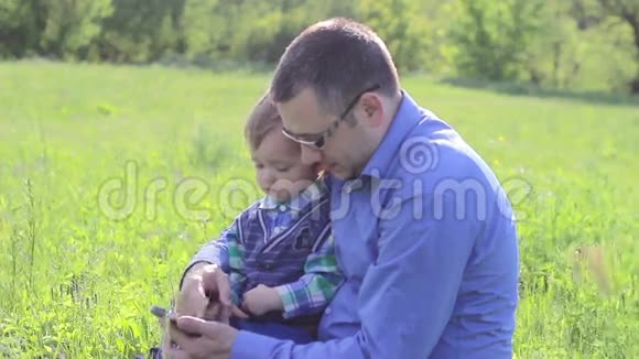 爸爸和儿子坐在草地上玩电话视频的预览图