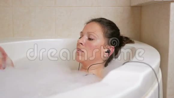 美丽的女孩洗澡泡沫和听音乐耳机一个大的白色浴缸和一杯红酒视频的预览图